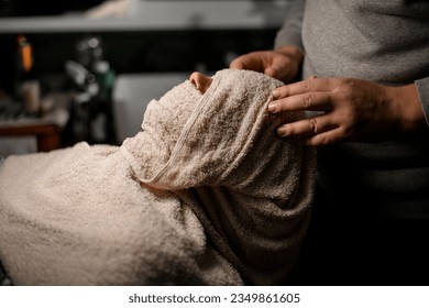 Hands of barber gently applying hot towel to male client's face to moisturize skin and beard hairs before shaving in modern barbershop - Powered by Shutterstock