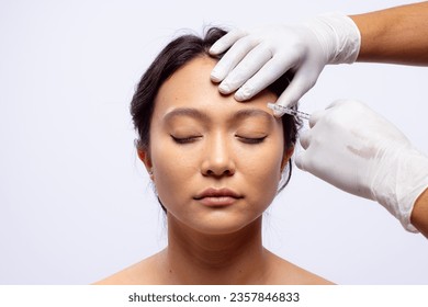 Hands of beautician in white surgical gloves injecting botox in brow of asian woman, copy space. Femininity, face, facial expressions, anti aging, skin and beauty treatments, unaltered.