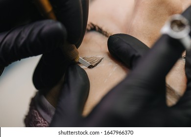 Hands in black gloves hold the manipulator with the microblading needle and draw her hair on the girl’s eyebrows close-up.