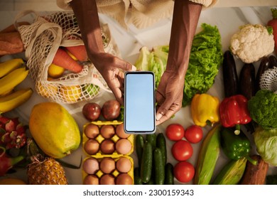 Hands of Black woman holding smartphone with empty screen over kitchen table with fresh fruits and vegetables - Powered by Shutterstock