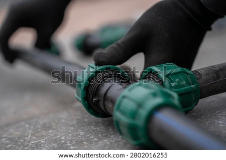 Hands in black work gloves assembling a plastic compression tee fitting connected to three HDPE pipes, installing a pipeline branch system for water supply or irrigation.