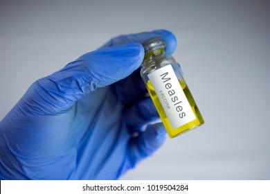 Hands In Blue Gloves Holding A Jar With A Yellow Vaccine. Measles Vaccination Concept. Close-up, Selective Focus