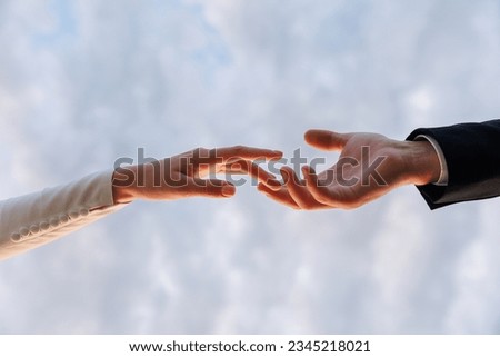 Hands of bride in white dress and groom in suit reaching each other, touching fingers on blue sky background. Helping hands for save and support people concept. Wedding day. Valentine day
