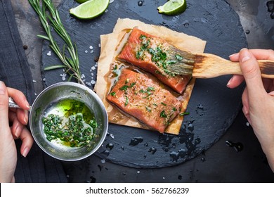 Hands Brush The Marinade Is Applied To A Piece Of Salmon On A Slate Stone On A Dark Metallic Background. View From Above. Preparation For Cooking Fish Food. Salmon Steak. Woman Cook.