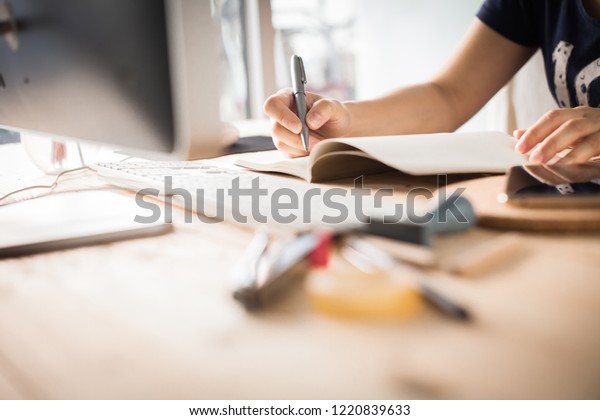 Hands of a Business woman is writing book as
notepad or Take notes on Her wooden table desk  . Business Success
Working at home office with notebook and computer for marketing and
part time Concept.