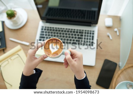 Hands of businesswoman holding cup of latte with heart shape foam after finishing working on laptop
