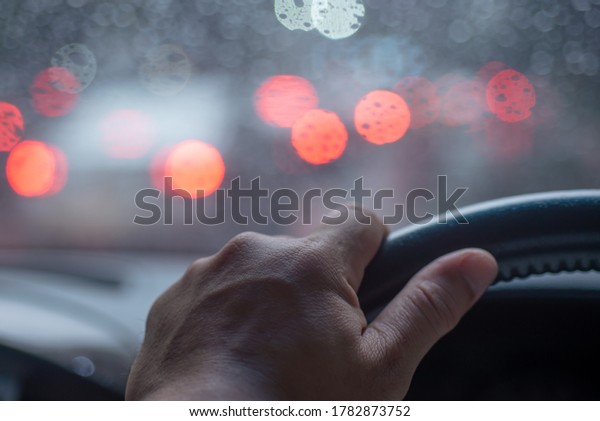 Hands of car driver on steering\
wheel, It s raining outside the windscreen, There are drops of\
water on the windscreen and blurred light, selective\
focus.