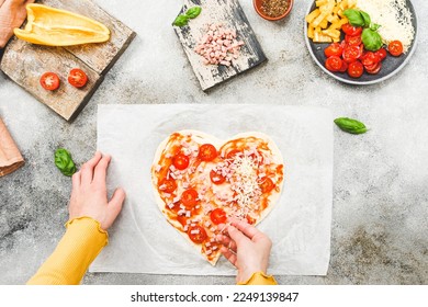 Hands of caucasian teenage girl lay down sliced cherry tomato on heart-shaped pizza dough for valentine's day with ingredients on the table, close-up side view. Valentine's day pizza making concept. - Powered by Shutterstock