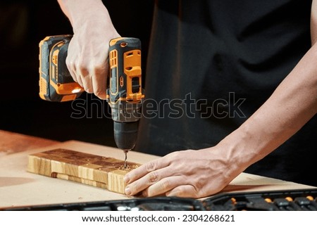 Hands of caucasian worker drilling a wooden detail with a cordless electric drill-driver on a wooden workbench