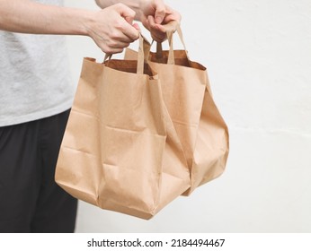 The Hands Of A Caucasian Young Man In A Gray T-shirt And Black Sweatpants Hold Out Two Paper Craft Bags With Fastwood Inside Against The Background Of A White Old Wall In The Backyard Of The House, 