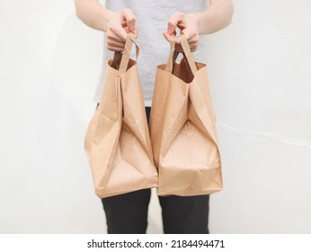 The Hands Of A Caucasian Young Man In A Gray T-shirt And Black Sweatpants Hold Out Two Paper Craft Bags With Fastwood Inside Against The Background Of A White Old Wall In The Backyard Of The House, 