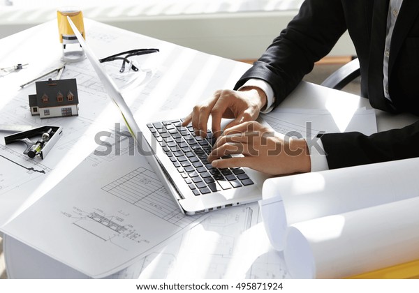 Hands of CEO of construction company in black suit\
typing message via e-mail to partners on laptop with drawings, seal\
stamp, divider on table. Young architect using netbook sitting in\
his office