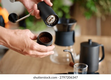Hands checking ground coffee in hand grinder, holding the opened grinder near a pour over coffee setup. Concept of coffee preparation and manual brewing.