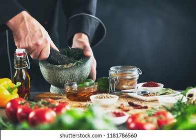 Hands of a chef grinding herbs in a pestle and mortar with assorted seasonings arranged on a wooden chopping board surrounded by fresh vegetables for salads - Powered by Shutterstock