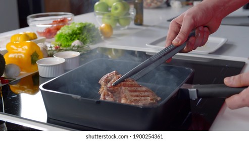 Hands Of Chef With Metallic Kitchen Tongs Turning Beef Steak On Hot Grill Frying Pan. Close Up Of Man Cooking Roasted Meat Standing By Electric Stove In Kitchen