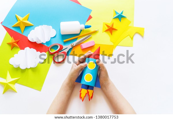hands of the child make a\
paper craft rocket. Cosmos clouds and stars colored paper. The\
creative process. Kindergarten and school development. Master\
Class.