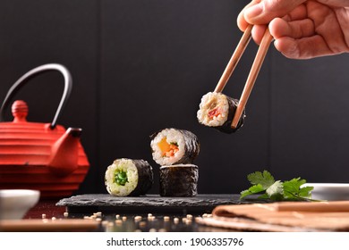 Hands with chopsticks picking sushi from slate plate with assortment on table with ingredients and containers and dark background. Front view. Horizontal composition. - Powered by Shutterstock