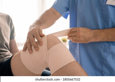 Hands Of Clinician In Blue Uniform Wrapping Injured Knee Of Patient With Flexible Bandage Before Rehabilitation Physical Training In Clinics