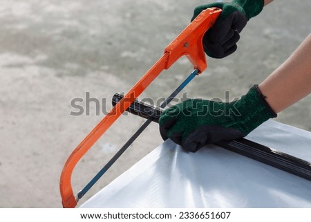 Hands of a construction worker wearing gloves is using a hacksaw to cut a piece of aluminum pipe.