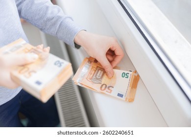 Hands counting euro banknotes while placing them on a radiator, symbolizing the rising utility costs and financial burden of energy bills during colder months.