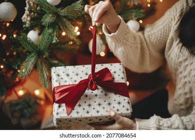 Hands in cozy sweater opening christmas gift with red bow on background of christmas tree with lights. Stylish female holding present with red ribbon in festive room close up. Merry Christmas! - Powered by Shutterstock