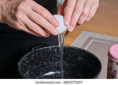Hands cracking egg into pot, clear yolk and white falling into dark pan, shell halves visible, focused wrist motion, kitchen mat under bowl, careful ingredient addition for batter or custard