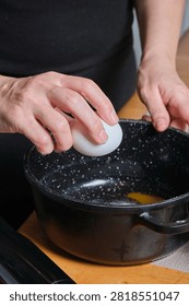 Hands cracking egg into pot, clear yolk and white falling into dark pan, shell halves visible, focused wrist motion, kitchen mat under bowl, careful ingredient addition for batter or custard