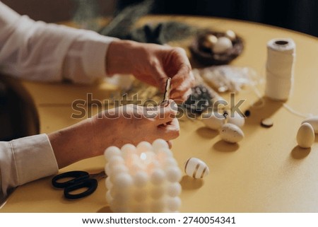 Hands crafting white eggs with string on a tabletop with scissors, thread spool and foam cube; cozy diy decorating workspace, shallow depth of field. Hands crafting decorative eggs with thread tools
