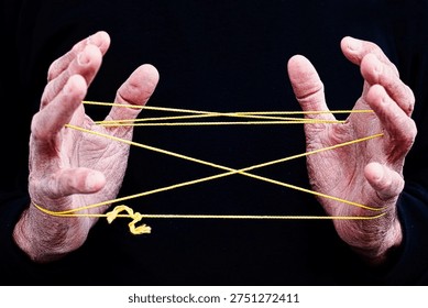 Hands Creating Cat’s Cradle String Figure on Black BackgroundThis image captures two hands skillfully creating a classic cat’s cradle string figure using a bright yellow string against a deep black background. The contrast highlights the intricate geometric pattern formed by the interwoven loops, showcasing the precision and coordination required for this traditional hand game. Cat’s cradle is a timeless activity known across many cultures, often passed down through generations as a simple yet engaging form of entertainment. 