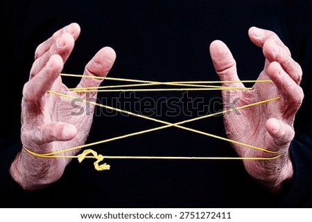 Hands Creating Cat’s Cradle String Figure on Black BackgroundThis image captures two hands skillfully creating a classic cat’s cradle string figure using a bright yellow string against a deep black background. The contrast highlights the intricate geometric pattern formed by the interwoven loops, showcasing the precision and coordination required for this traditional hand game. Cat’s cradle is a timeless activity known across many cultures, often passed down through generations as a simple yet engaging form of entertainment. 
