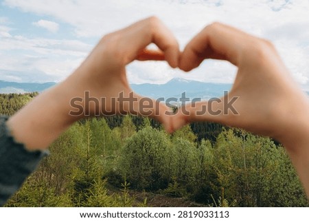 Hands creating a heart shape, framing a scenic mountain range with green forests and a distant snowy peak under cloudy sky