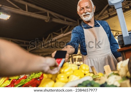 Hands of a customer and a vendor making transaction with a credit card and a pos terminal. A smiling seller charges for his vegetables at the stand.