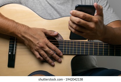 Hands of a dark man on the six strings of a guitar looking at his cell phone looking for new music to play in his home. Wearing a gray t-shirt on a white wall. - Powered by Shutterstock