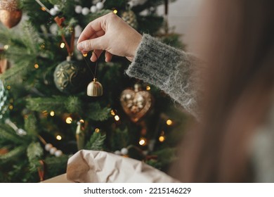 Hands decorating christmas tree with stylish bauble in atmospheric festive room. Merry Christmas! Winter holidays preparation. Woman in cozy sweater putting vintage bell on tree