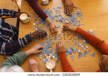 Hands of diverse group of senior friends playing with jigsaw puzzles in sunny dining room at home. Retirement, friendship, wellbeing, activities, togetherness and senior lifestyle, unaltered.