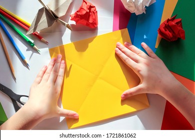 Children’s hands doing origami crane from yellow paper on white background with various school supplies. Step-by-step tutorial of origami. Step 4. Concept of children's creativity, back to school.