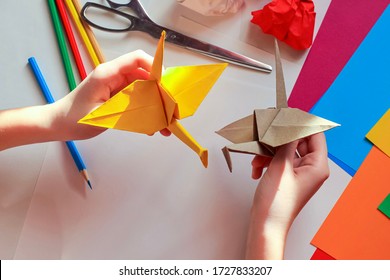 Children’s hands doing origami crane from yellow paper on white background with various school supplies. Step-by-step tutorial of origami. Step 18. Concept of children's creativity, back to school.