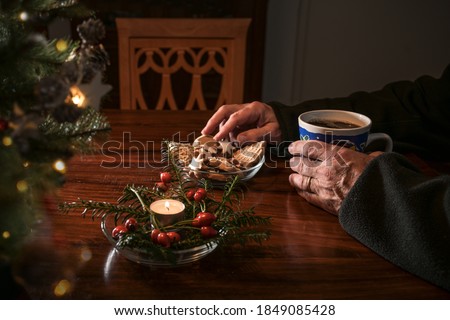 Hands of an elderly lonely man on a table with Christmas cookies, coffee and festive decoration next to an empty chair, sad holidays during the croronavirus pandemic or after a loss