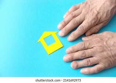 Hands Of An Elderly Man And A House Cut Out Of Colored Paper On A Blue Background, Copyspace On The Left. The Concept Of A Nursing Home, Lonely Old Age, Real Estate Purchase, Home Insurance.