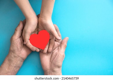 The hands of an elderly person and a child hold a red heart embedded in paper. Caring for the health of the heart of the elderly. Right copyspace. Love for old relatives concept - Shutterstock ID 2140465739