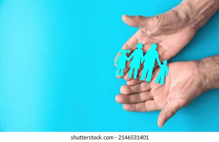 The Hands Of An Elderly Person Hold A Paper-cut Silhouette Of A Family On A Blue Fog. Copyspace On The Left. Family Day, Family Traditions Concept. Family Tree, Succession Of Generations.