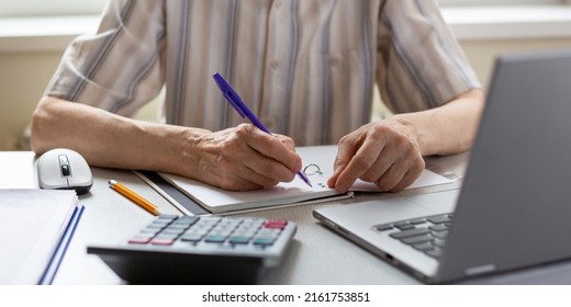 Hands Of Elderly Person. Senior Woman Writes Down Something Sitting At The Laptop At Home, Selective Focus. Study, Audit, Remote Work, Accountant, Calculator, Count, Tutor, WFH, Retraining Concept