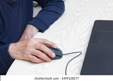 Hands Of An Elderly Woman Close-up With A Computer Mouse. A Woman In A Blue Sweater Sits At A Table And Works On A Laptop. The Concept Of Retraining Of Older People, Computer Training