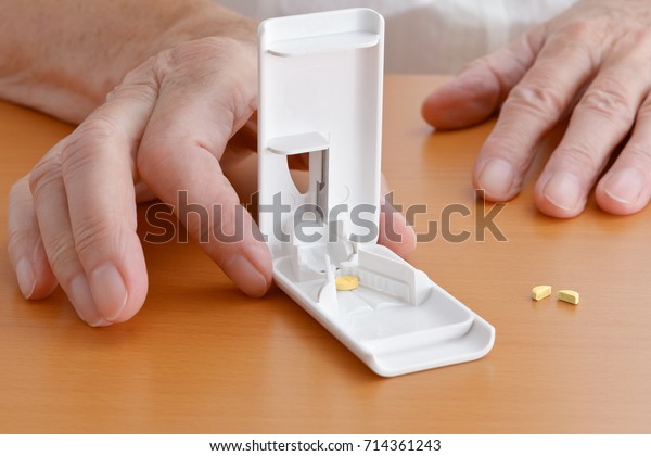 Hands of an elderly woman on a wooden tabletop\
holding a white plastic pill or tablet cutter, one pill inside, one\
divided in halves beside