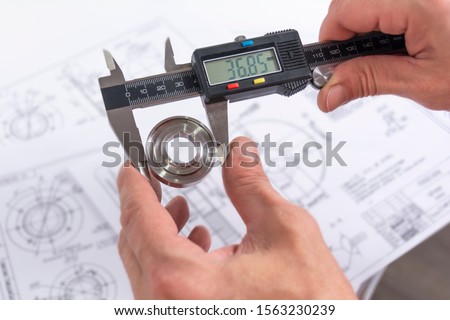 Hands of an engineer measures a metal part with a digital vernier caliper against the background of technical drawings. Quality control of parts machined on a lathe.