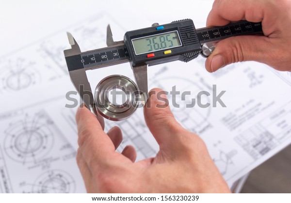 Hands of an engineer measures\
a metal part with a digital vernier caliper against the background\
of technical drawings. Quality control of parts machined on a\
lathe.