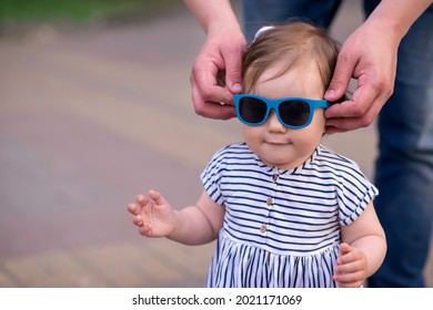 Hands Of Father Putting Sunglasses On Little Daughter During Walk. Close Up Photo: Dad Puts Shades On Smiling Toddler Girl In Striped Dress. Parental Care, Eye Protection, Summer And Vacation Concept