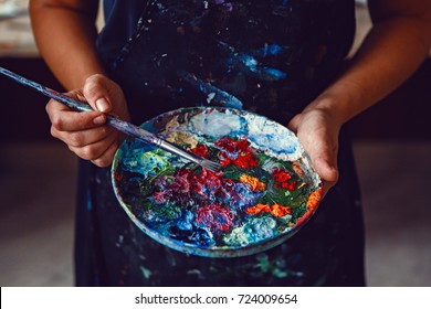 Hands Of Female Artist Holding Messy Dirty Palette  With Different Paints And Paintbrush In Art Studio. Lifestyle And Hobby Concept 