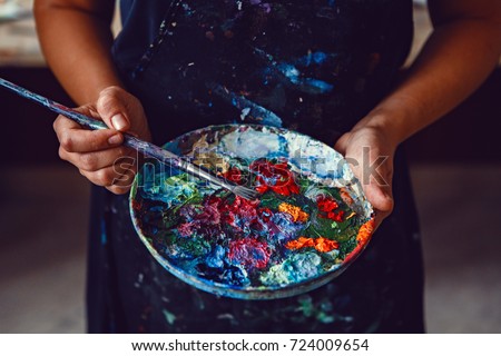 Hands of female artist holding messy dirty palette  with different paints and paintbrush in art studio. Lifestyle and hobby concept 