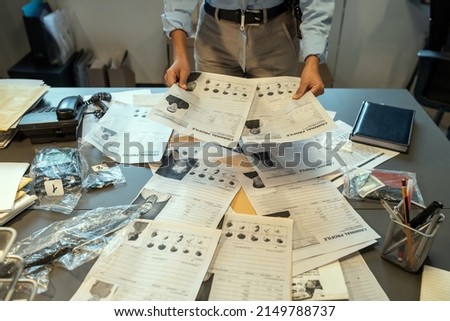 Hands of female investigator holding criminal profiles over desk with documents while learning personal information about suspects